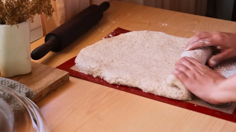 formado-y-enrollado-pan-integral-coffee-maria Manos de panadera enrollando cuidadosamente una masa de pan integral extendida sobre un tapete de silicona rojo para darle forma de cilindro.