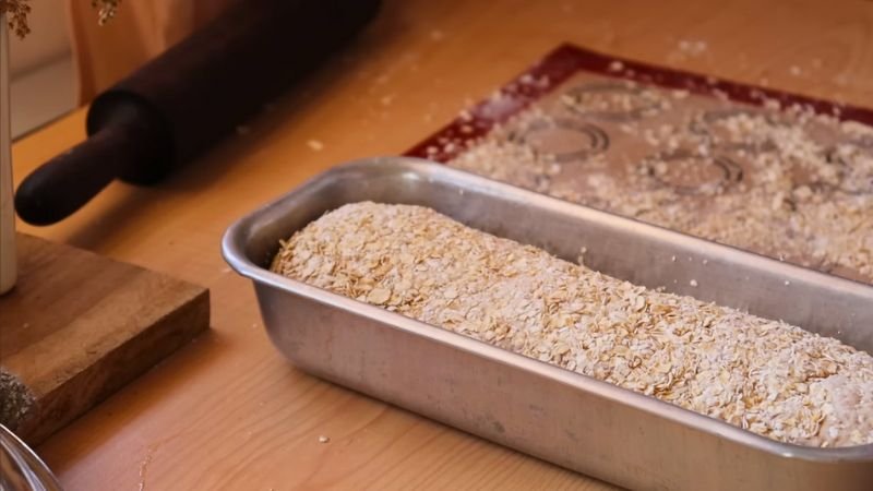 pan-integral-con-avena-en-molde Masa de pan integral enrollada y cubierta con hojuelas de avena dentro de un molde metálico rectangular sobre una mesa de madera.