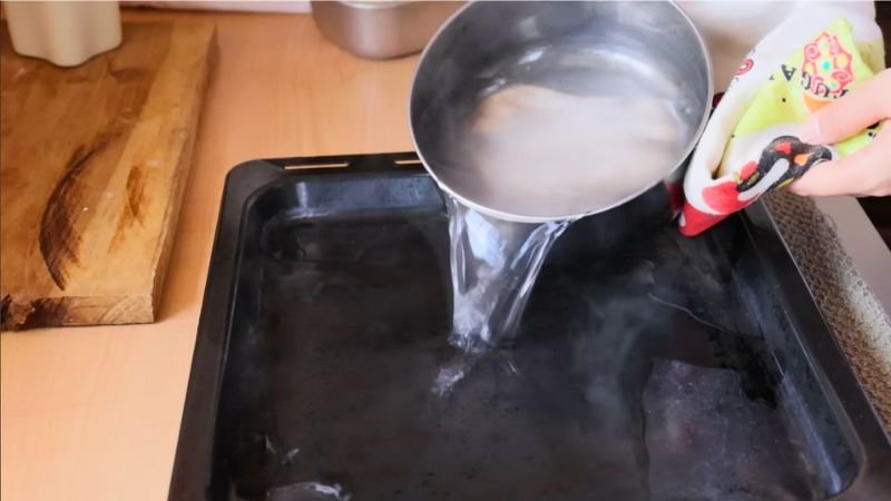 tecnica-vapor-horno-corteza-crujiente Acción de verter agua caliente desde un cazo hacia una bandeja negra de horno para crear vapor durante el horneado del pan integral de masa madre fácil y crujiente