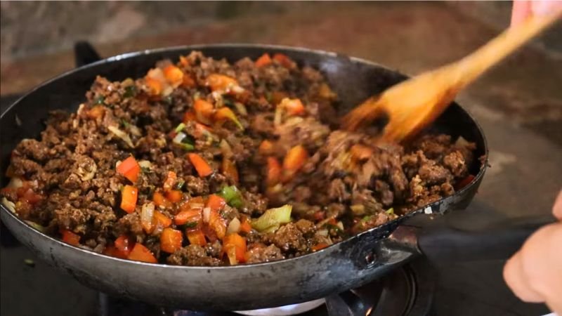 Integración de sabores para el relleno - Coffee María. Sartén con carne molida mezclada con trozos de tomate, pimentón y cebollín siendo removidos con una cuchara de madera.