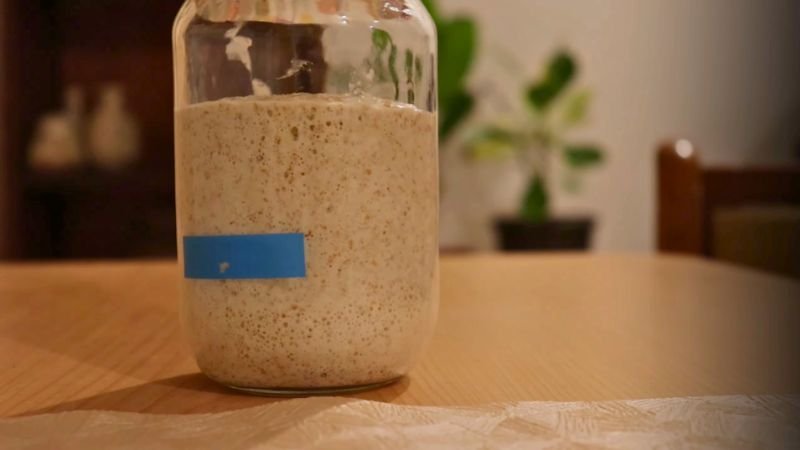 Captura del video de Coffee Maria mostrando el descarte de la mitad y la adición de 100g de harina integral y 50g de agua en el día 3.