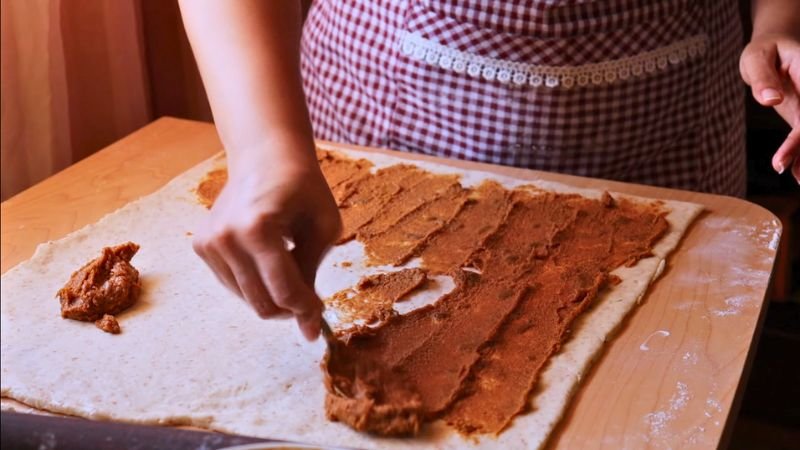 Técnica de Relleno y Formado de los Roles. Vista superior de una persona usando un delantal de cuadros mientras extiende una pasta cremosa de canela y papelón sobre una lámina rectangular de masa flexible para preparar roles de masa madre artesanales.