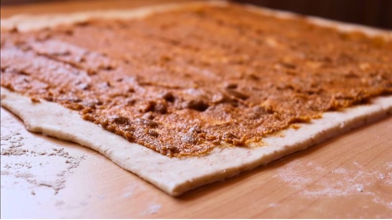 Masa Estirada con Relleno de Canela y Papelón. Un primer plano en plano picado que muestra una lámina rectangular de masa flexible completamente estirada sobre una tabla de madera, cubierta uniformemente con una pasta cremosa de canela y papelón para preparar roles de masa madre artesanales.