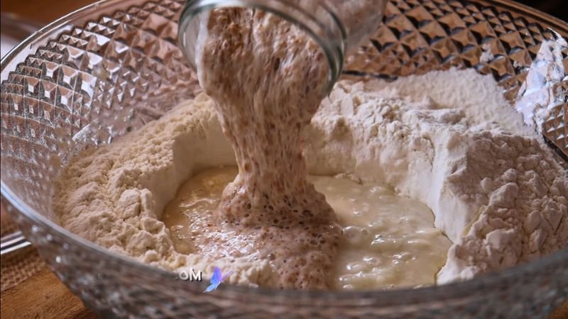 Activación de la Masa Madre Burbujeante. Un primer plano de un tazón de vidrio grande y texturizado, donde una mano vierte una masa madre burbujeante y activa desde un frasco de vidrio sobre una mezcla de harina panadera, agua y azúcar, iniciando el proceso de los roles de masa madre de Coffee María.