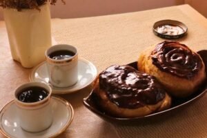 Una toma de primer plano con luz natural que muestra dos roles de canela de masa madre cubiertos de chocolate brillante en un cuenco marrón, acompañados de dos tazas de café sobre un mantel rústico en Coffee María.
