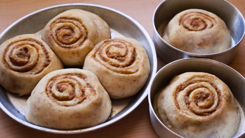 Roles de Masa Madre Formados en Bandejas. Un plano picado muestra varias bandejas de aluminio circulares sobre una superficie de madera, cada una conteniendo roles de canela de masa madre recién cortados y formados. Se aprecian las espirales de masa y relleno de canela antes de su fermentación final y horneado.