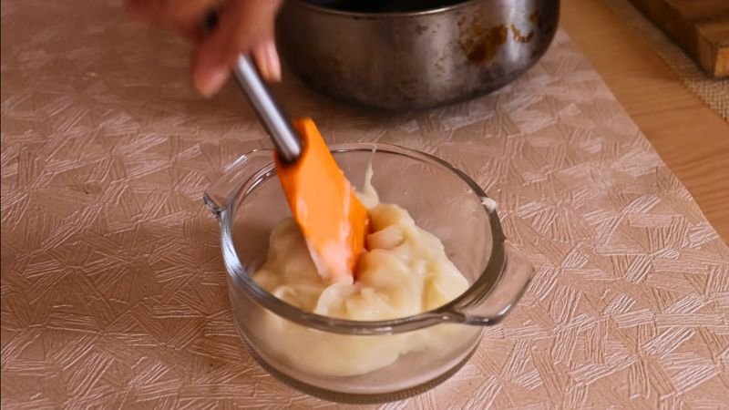 Textura Gelatinosa del Tangzhong. Una toma de primer plano muestra una mano usando una espátula de cocina naranja para revolver la pasta de harina y agua cocida (Tangzhong) en un tazón de vidrio pequeño, asegurando que la textura sea suave y gelatinosa para los roles de masa madre.