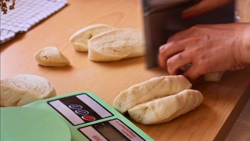 Cortando y pesando porciones de masa de pan francés con una balanza digital verde sobre un mesón de madera.