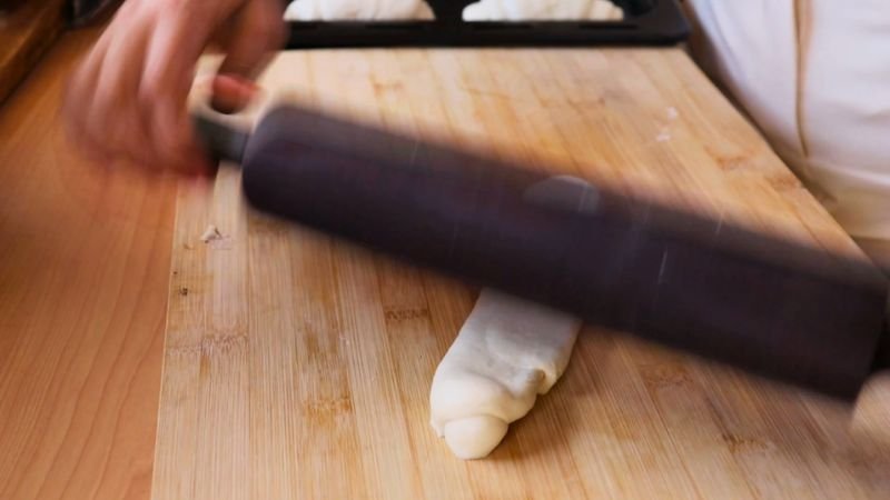 Manos utilizando un rodillo negro para aplanar una porción de masa de pan francés sobre un mesón de madera.
