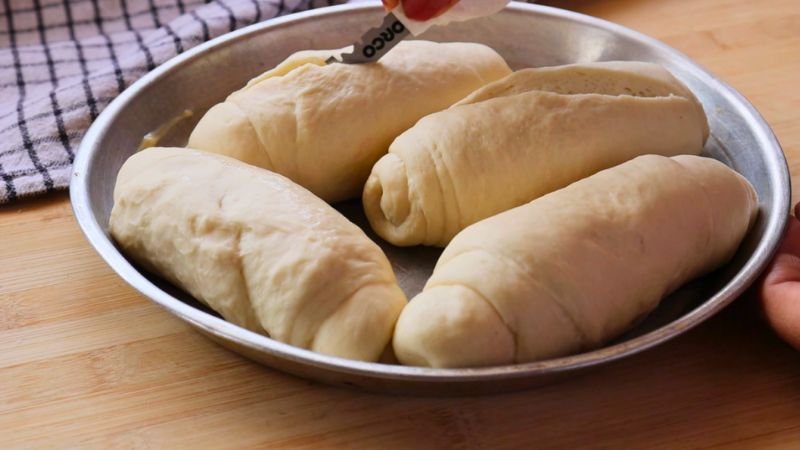 Cuatro panes de masa madre colocados en una bandeja redonda de aluminio, con una hojilla a punto de realizar un corte longitudinal.