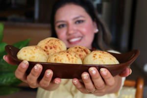 María sonriendo mientras sostiene un plato marrón con pan de queso brasileño (pão de queijo tradicional) recién horneado.