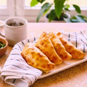 Cuatro empanadas doradas sin gluten servidas en una bandeja con paño de cocina, acompañadas de una taza de café caliente con vapor.