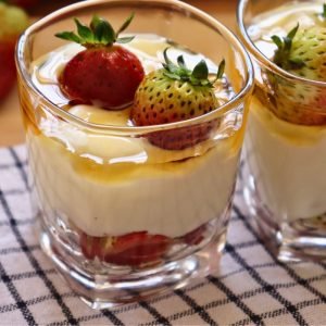 Vaso de cristal con capas de fresas frescas, crema blanca suave y un toque de miel o almíbar en la parte superior, servido sobre un mantel a cuadros en Coffee Maria.