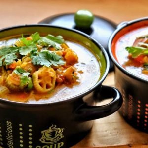 Tazón negro de sopa de vegetales con cilantro fresco y rodajas de quimbombó (okra), servido en una mesa de madera.